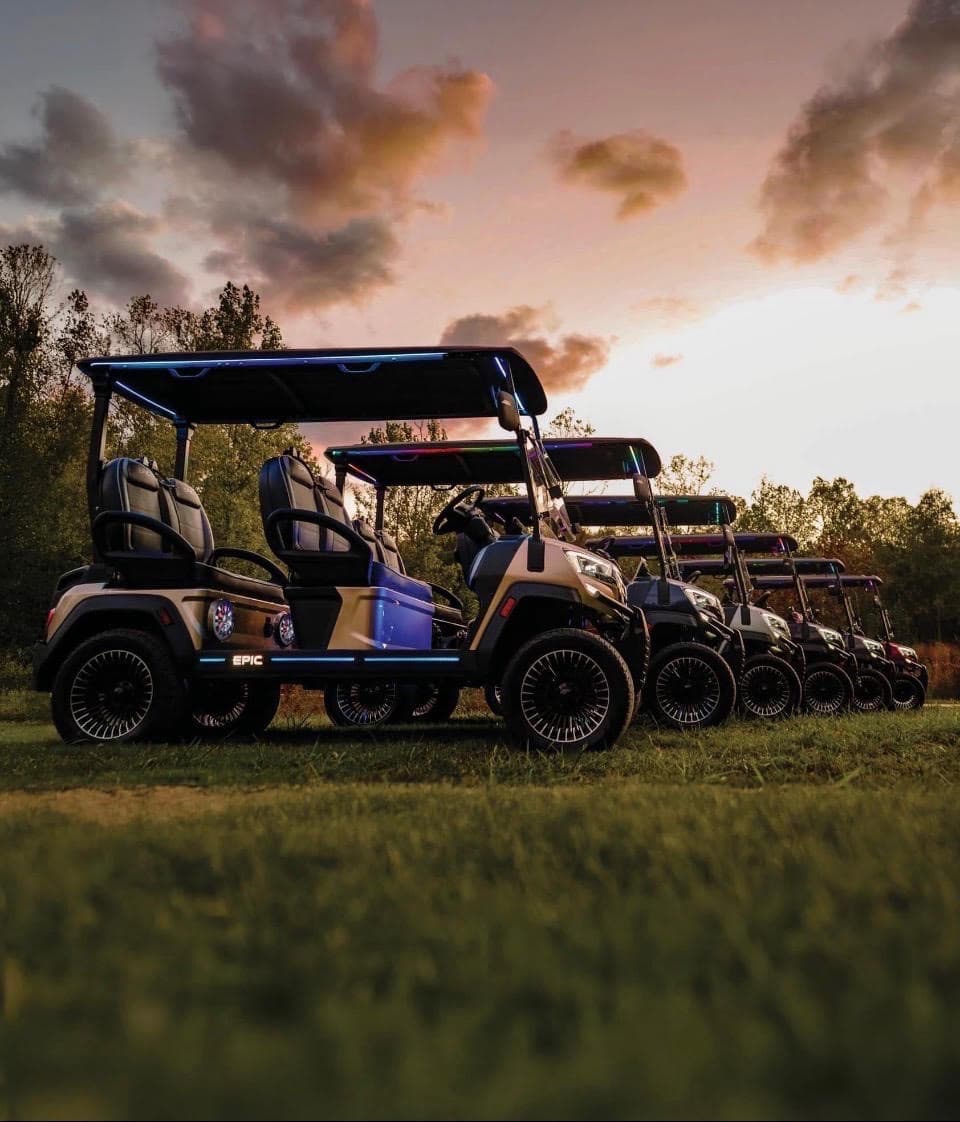 Row of EPIC golf carts lined up on grass at sunset with glowing underbody lights and a pink and orange sky